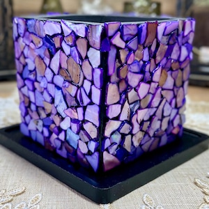 May include: A square glass candle holder with a purple mosaic design. The holder is made of small, irregular pieces of glass that are glued together to create a unique pattern. The holder is sitting on a black base.