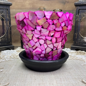 May include: A decorative planter with a mosaic design made of pink and purple shell pieces. The planter sits on a black saucer. Two black candle lanterns are in the background.
