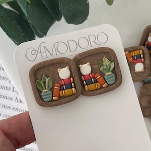 May include: A pair of square-shaped earrings featuring a brown border with a white bear wearing a colorful backpack and a small potted plant. The earrings are on a white card with the brand name "AMODORO". Additional earrings are visible.