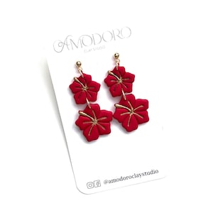 May include: Red polymer clay earrings with gold accents, featuring two hibiscus flowers on each earring. The earrings are displayed on a white card with the text "AMODORO CLAY STUDIO" and "@amodoroclaystudio".