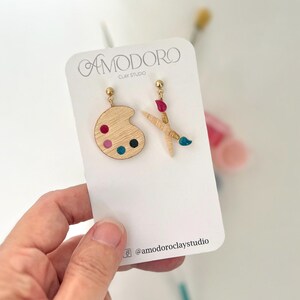 May include: Wooden artist-themed earrings. One earring is a palette with colorful paint dots, the other is crossed paintbrushes with red and blue paint. The earrings are on a white card with the text "AMODORO CLAY STUDIO".