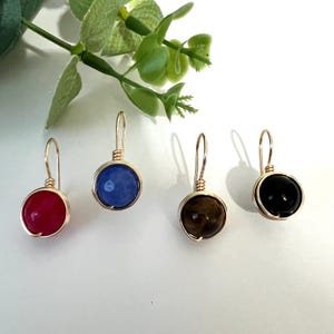 May include: Four wire-wrapped dangle earrings featuring round beads in red, blue, brown, and black. Each earring is suspended from a gold-toned metal hook and secured with a coiled wire setting that encircles the circumference of the gemstone bead.