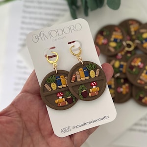 May include: A pair of handcrafted clay earrings featuring a miniature bookshelf design. Each earring showcases a brown circular shelf with tiny books, plants, and a mushroom. The earrings hang from gold-colored hoops. The brand name "AMODORO CLAY STUDIO" is visible.