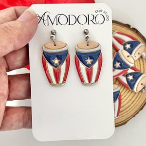May include: A pair of dangle earrings on a white display card labeled AMODORO CLAY STUDIO. The earrings are shaped like drums with a tan top, a blue upper section with a white star, and vertical red and white stripes on the lower body.