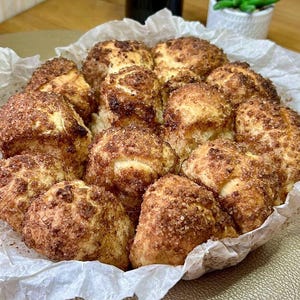 May include: A close-up shot of a baked dessert, likely cinnamon rolls, arranged in a white paper-lined basket. The rolls are golden brown with a generous coating of cinnamon sugar. The background includes small potted plants.