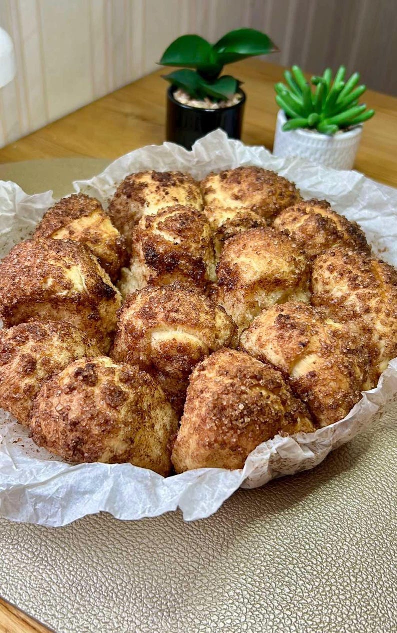 May include: A close-up shot of a baked dessert, likely cinnamon rolls, arranged in a white paper-lined basket. The rolls are golden brown with a generous coating of cinnamon sugar. The background includes small potted plants.