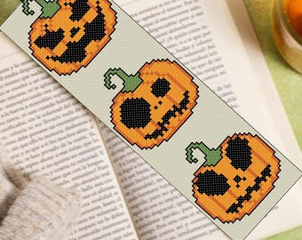 May include: A beige bookmark with three orange and black pixelated jack-o'-lantern designs. The bookmark is on top of an open book.