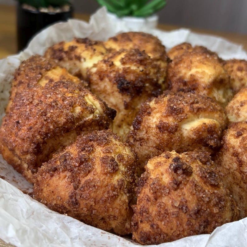May include: A close-up shot of a basket filled with freshly baked cinnamon rolls. The rolls are golden brown with a generous coating of cinnamon sugar. The rolls are arranged in a white paper-lined basket, ready to be enjoyed.