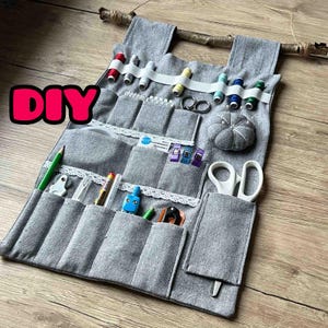 Puede incluir: Organizador de tela gris DIY con múltiples bolsillos, colgado de una rama. El organizador está lleno de artículos de costura, incluyendo carretes de hilo, tijeras, agujas y un alfiletero.