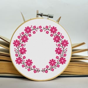 May include: A pink floral wreath cross stitch pattern in a wooden embroidery hoop.