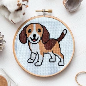 Può includere: Un ricamo a punto croce di un beagle in un cerchio di legno rotondo. Il beagle è marrone, bianco e nero con un'espressione felice.