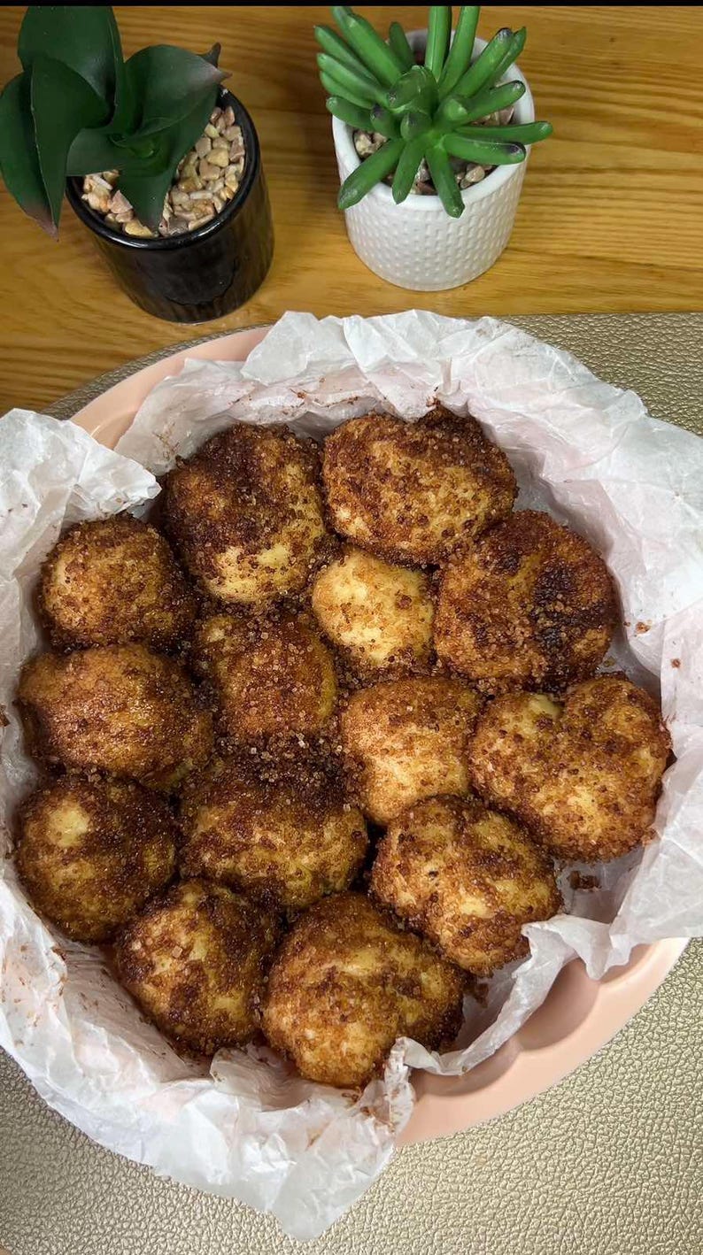May include: A close-up shot of a pink plate filled with golden-brown, round pastries. The pastries are coated in a dark brown sugar and cinnamon mixture, sitting on white parchment paper. The image suggests a dessert or snack.