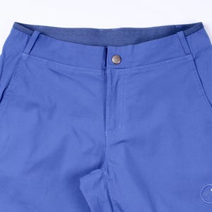 Puede incluir: Primer plano de unos pantalones cortos azules con cierre de bot&oacute;n y bolsillos laterales. Los pantalones cortos tienen una cinturilla azul oscuro y un peque&ntilde;o logotipo en la parte inferior derecha. La tela parece ser un material duradero y ligero.