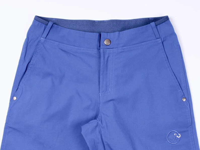 Puede incluir: Primer plano de unos pantalones cortos azules con cierre de bot&oacute;n y bolsillos laterales. Los pantalones cortos tienen una cinturilla azul oscuro y un peque&ntilde;o logotipo en la parte inferior derecha. La tela parece ser un material duradero y ligero.