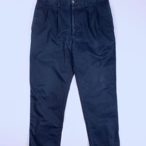 May include: Dark navy blue trousers with a button fastening and pleats. These straight-leg trousers are made from a solid, dark blue material. The trousers are shown against a white backdrop.