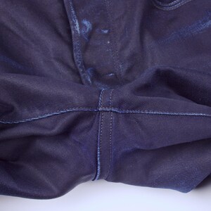 May include: Close-up of dark blue denim jeans. The image highlights the crotch area, showcasing the stitching and fabric texture. The denim appears to have subtle fading and a slightly worn appearance.