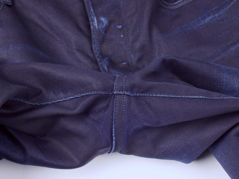 May include: Close-up of dark blue denim jeans. The image highlights the crotch area, showcasing the stitching and fabric texture. The denim appears to have subtle fading and a slightly worn appearance.