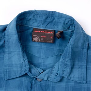 Puede incluir: Una camisa de cuadros azul con cuello en punta. Una etiqueta interior marr&oacute;n oscuro muestra el logotipo de Mammut, el texto Mammut Swiss Design e informaci&oacute;n de tallas que incluye Europe M, Asia L y China 175/92B. La tela tiene un patr&oacute;n de rejilla.