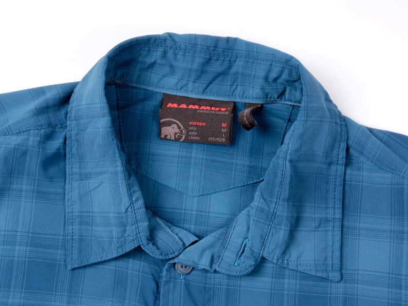 Puede incluir: Una camisa de cuadros azul con cuello en punta. Una etiqueta interior marr&oacute;n oscuro muestra el logotipo de Mammut, el texto Mammut Swiss Design e informaci&oacute;n de tallas que incluye Europe M, Asia L y China 175/92B. La tela tiene un patr&oacute;n de rejilla.