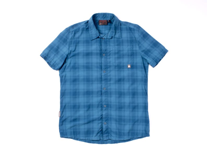 Puede incluir: Una camisa de manga corta con botones en un patr&oacute;n de cuadros azules. La camisa presenta un cuello est&aacute;ndar, un bolsillo en el pecho en el lado izquierdo con una peque&ntilde;a etiqueta rectangular con el logotipo y una fila de botones azules.