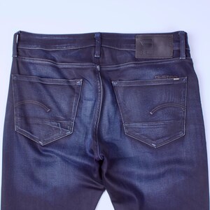 May include: Dark blue denim jeans, rear view. Features include two back pockets with curved stitching, a leather patch at the waistband, and the brand name "G-Star" visible. The jeans have a classic five-pocket design.