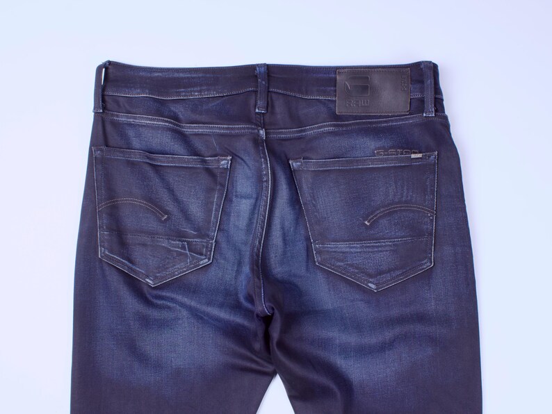 May include: Dark blue denim jeans, rear view. Features include two back pockets with curved stitching, a leather patch at the waistband, and the brand name "G-Star" visible. The jeans have a classic five-pocket design.