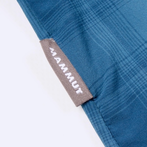 Puede incluir: Una prenda de tela azul con un sutil patr&oacute;n de cuadros y una peque&ntilde;a etiqueta rectangular de color marr&oacute;n claro cosida en el borde. La etiqueta muestra la palabra MAMMUT en letras blancas, may&uacute;sculas y sin remates, orientada verticalmente.