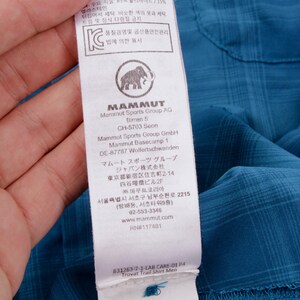 Puede incluir: Una camisa de cuadros azul con una etiqueta de cuidado blanca en el interior que muestra el logotipo de Mammut, un icono de mamut y texto impreso que incluye Mammut Sports Group AG, direcciones, sitio web y Trovat Trail Shirt Men.