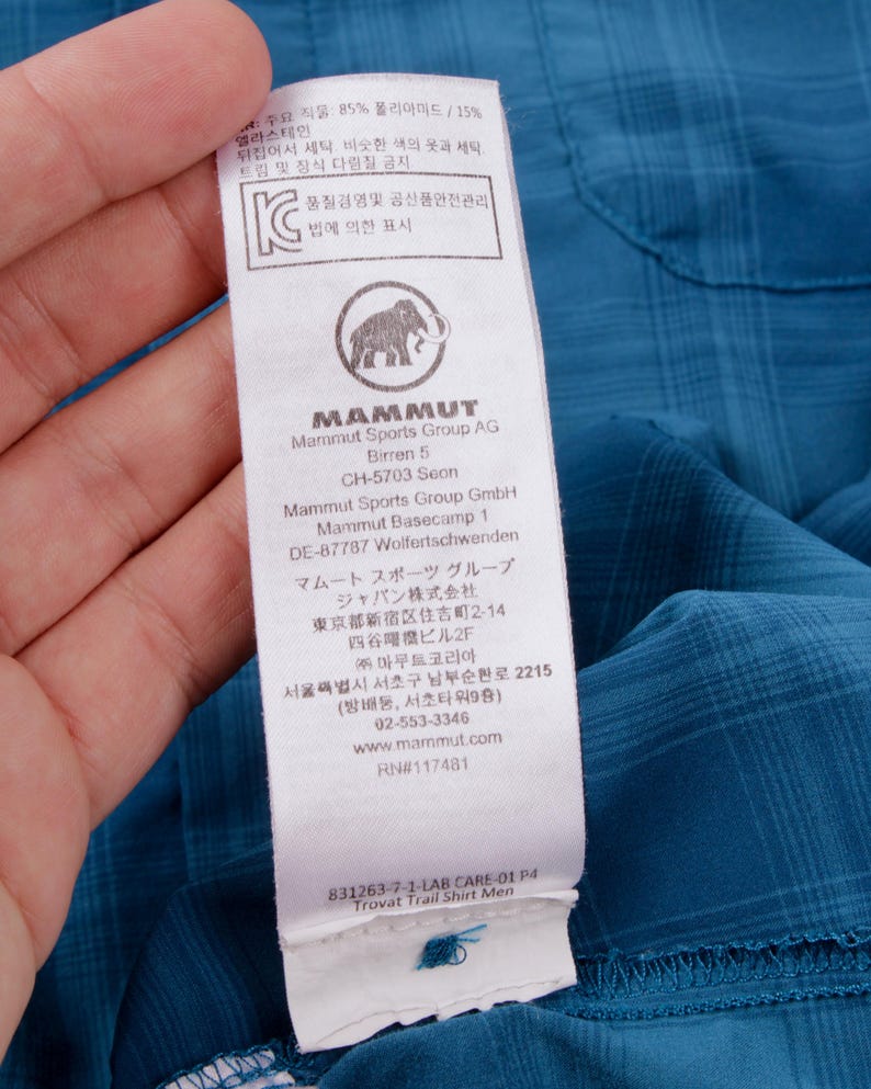 Puede incluir: Una camisa de cuadros azul con una etiqueta de cuidado blanca en el interior que muestra el logotipo de Mammut, un icono de mamut y texto impreso que incluye Mammut Sports Group AG, direcciones, sitio web y Trovat Trail Shirt Men.