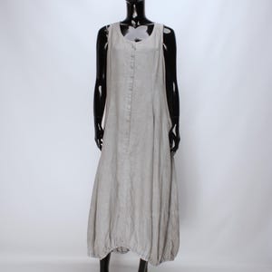 Può includere: Abito in lino grigio chiaro su un manichino nero. L'abito senza maniche ha un'abbottonatura frontale e un orlo a palloncino arricciato. L'abito è di colore neutro e sembra essere realizzato in un tessuto leggero.