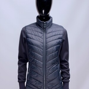 Può includere: Un gilet trapuntato blu navy con chiusura a zip completa e collo alto. Il gilet presenta un motivo a spina di pesce ed è abbinato a maniche lunghe. Il manichino indossa pantaloni bianchi.