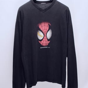 May include: Black long-sleeve V-neck shirt featuring a Spider-Man mask graphic. The mask is red with white eye shapes and a web design. The shirt has the text "SENSATIONAL TOUR" and is hanging on a black hanger.