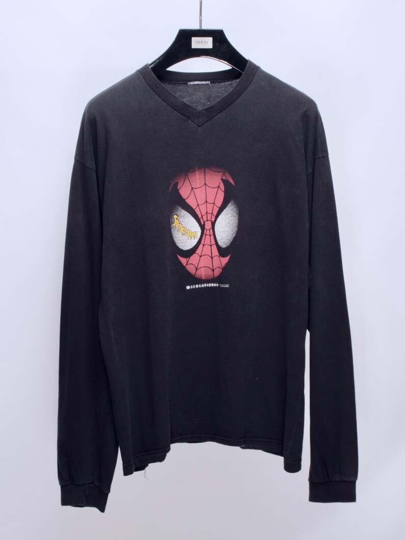 May include: Black long-sleeve V-neck shirt featuring a Spider-Man mask graphic. The mask is red with white eye shapes and a web design. The shirt has the text "SENSATIONAL TOUR" and is hanging on a black hanger.