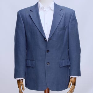 Puede incluir: Un blazer azul con una camisa blanca, exhibido en un maniquí de madera. El blazer tiene dos bolsillos delanteros y un cierre de dos botones. Las mangas son largas y el cuello de la camisa es visible.