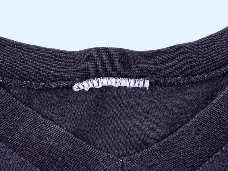 May include: Close-up of a dark gray V-neck shirt. The neckline and collar are ribbed, and a white tag is sewn into the back of the collar. The fabric has a slightly worn appearance.
