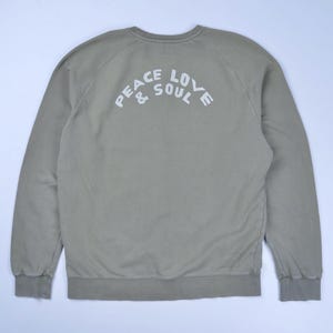 May include: A sage green sweatshirt featuring the white text "PEACE LOVE & SOUL" arched across the upper back. The long-sleeved top has a crew neck and ribbed cuffs and hem.