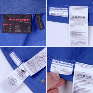 Puede incluir: Primer plano de tela azul con varias etiquetas. Las etiquetas muestran informaci&oacute;n de tallas, n&uacute;meros de estilo e instrucciones de cuidado. La marca "MAMMUT" es visible en una etiqueta. La tela parece ser parte de una prenda.