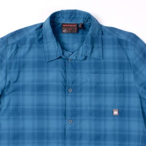 Puede incluir: Una camisa de manga corta con botones y un patr&oacute;n de cuadros azules. La camisa cuenta con un cuello est&aacute;ndar, una tapeta de botones frontal y un bolsillo en el pecho en el lado izquierdo con una peque&ntilde;a etiqueta de la marca Mammut en la esquina.
