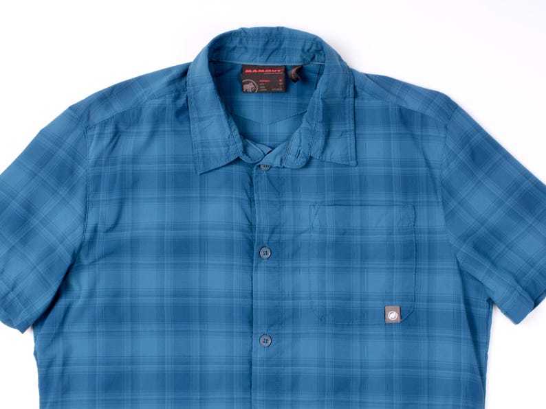 Puede incluir: Una camisa de manga corta con botones y un patr&oacute;n de cuadros azules. La camisa cuenta con un cuello est&aacute;ndar, una tapeta de botones frontal y un bolsillo en el pecho en el lado izquierdo con una peque&ntilde;a etiqueta de la marca Mammut en la esquina.