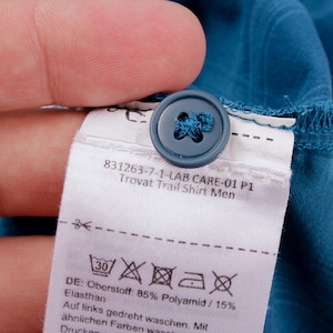 Puede incluir: Una camisa de color azul cerceta con un bot&oacute;n a juego y una etiqueta de cuidado blanca con el texto 831263-7-1-LAB CARE-01 P1 Trovat Trail Shirt Men, s&iacute;mbolos de lavado y composici&oacute;n de tela de 85% Polyamid y 15% Elasthan.
