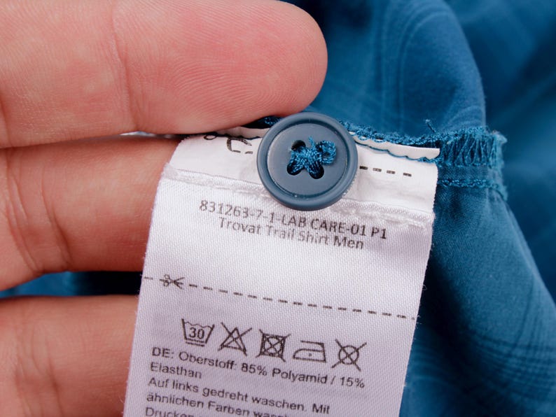 Puede incluir: Una camisa de color azul cerceta con un bot&oacute;n a juego y una etiqueta de cuidado blanca con el texto 831263-7-1-LAB CARE-01 P1 Trovat Trail Shirt Men, s&iacute;mbolos de lavado y composici&oacute;n de tela de 85% Polyamid y 15% Elasthan.