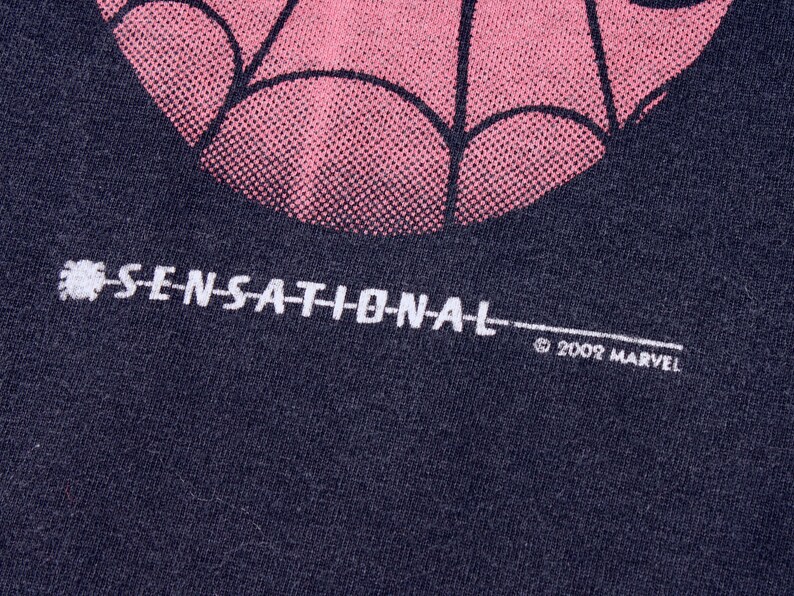 May include: Close-up of a dark blue t-shirt with a pink and black spiderweb design. The word "SENSATIONAL" is printed in white, along with the copyright "&copy; 2002 MARVEL".