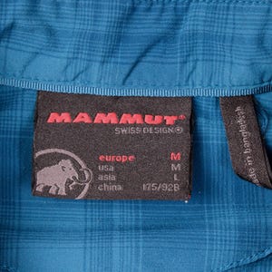 Puede incluir: Una camisa de cuadros azul con una etiqueta interior negra que muestra el logotipo rojo de Mammut, un gr&aacute;fico de mamut e informaci&oacute;n de tallas para Europa, EE. UU., Asia y China. Una etiqueta negra m&aacute;s peque&ntilde;a indica su fabricaci&oacute;n.
