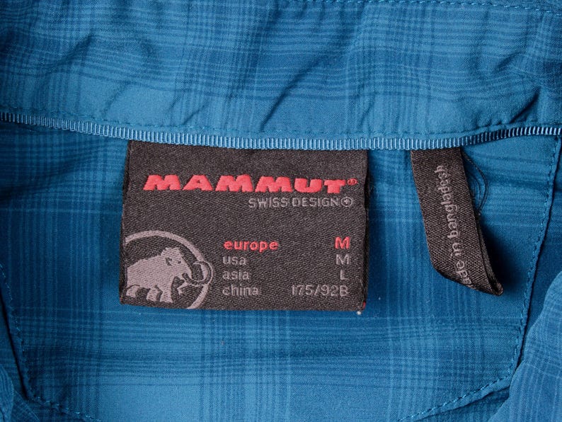 Puede incluir: Una camisa de cuadros azul con una etiqueta interior negra que muestra el logotipo rojo de Mammut, un gr&aacute;fico de mamut e informaci&oacute;n de tallas para Europa, EE. UU., Asia y China. Una etiqueta negra m&aacute;s peque&ntilde;a indica su fabricaci&oacute;n.