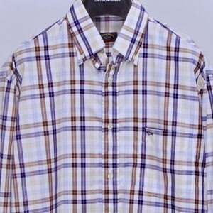 May include: A button-down shirt with a plaid pattern in white, brown, light blue, and purple. The shirt has a small pocket on the left chest and is hanging on a black hanger. The hanger has the text "EMPORIO ARMANI".