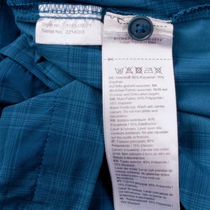 Puede incluir: Una camisa de cuadros color verde azulado con una etiqueta de cuidado interior blanca con el texto Style no. 1015-00071 y Serial No. 2214088. Un bot&oacute;n azul oscuro est&aacute; unido a la etiqueta, que indica 85% Poliamida y 15% Elastano.