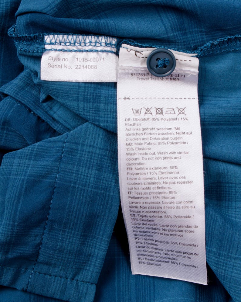 Puede incluir: Una camisa de cuadros color verde azulado con una etiqueta de cuidado interior blanca con el texto Style no. 1015-00071 y Serial No. 2214088. Un bot&oacute;n azul oscuro est&aacute; unido a la etiqueta, que indica 85% Poliamida y 15% Elastano.