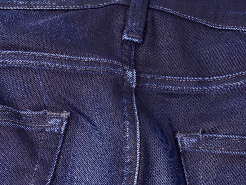 May include: Close-up of the back of dark blue denim jeans. The image highlights the details of the pockets, stitching, and belt loops. The denim has a slightly faded appearance, with visible wear and tear.