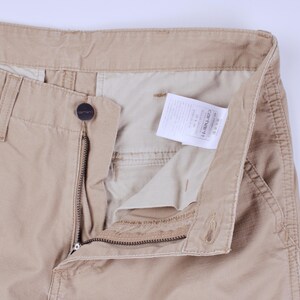 Pode incluir: Close-up de cal&ccedil;as Carhartt bege, parcialmente desabotoadas e com z&iacute;per aberto, revelando o forro interno e uma etiqueta branca. As cal&ccedil;as apresentam fecho de bot&atilde;o, z&iacute;per e presilhas para cinto. O logotipo da Carhartt &eacute; vis&iacute;vel.