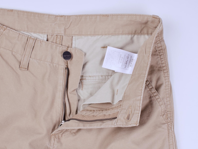 Pode incluir: Close-up de cal&ccedil;as Carhartt bege, parcialmente desabotoadas e com z&iacute;per aberto, revelando o forro interno e uma etiqueta branca. As cal&ccedil;as apresentam fecho de bot&atilde;o, z&iacute;per e presilhas para cinto. O logotipo da Carhartt &eacute; vis&iacute;vel.